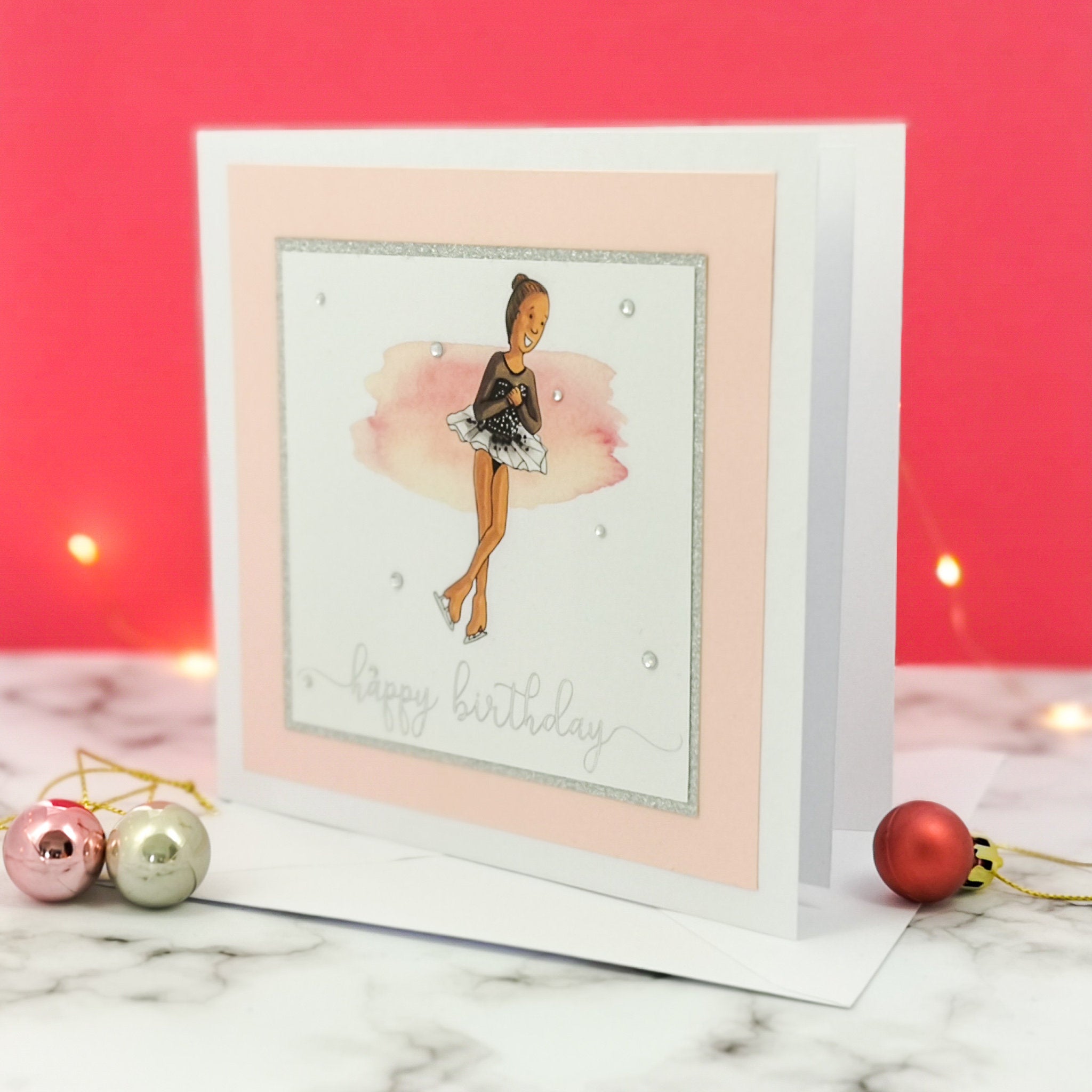 6 x 6 inch birthday card with image of a figure skater mid-flight, wearing a black dress. Pink watercolour brush stroke behind the figure skater illustration, with a silver foiled happy birthday script greeting and surrounded by clear/silver rhinestone gems