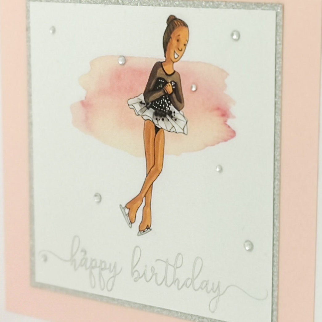 Close up of 6 x 6 inch birthday card with image of a figure skater mid-flight, wearing a black dress. Pink watercolour brush stroke behind the figure skater illustration, with a silver foiled happy birthday script greeting and surrounded by clear/silver rhinestone gems