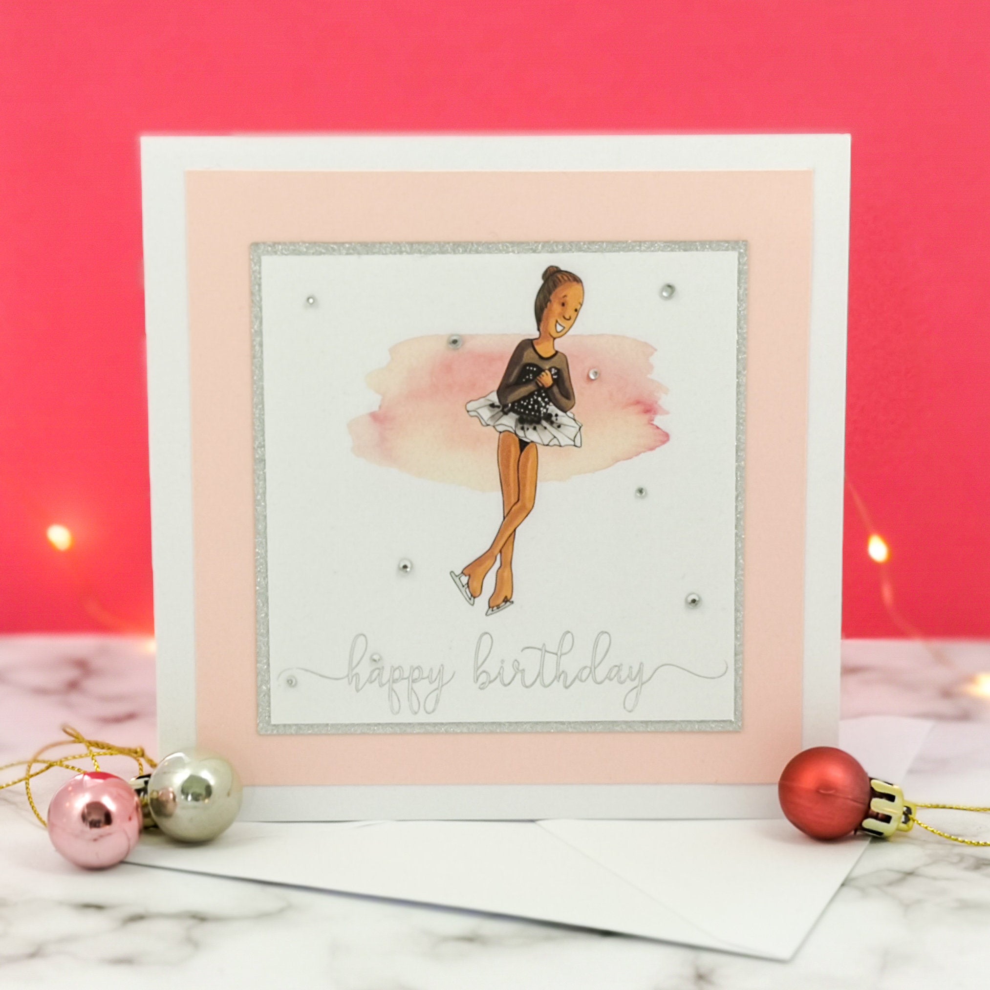 6 x 6 inch birthday card with image of a figure skater mid-flight, wearing a black dress. Pink watercolour brush stroke behind the figure skater illustration, with a silver foiled happy birthday script greeting and surrounded by clear/silver rhinestone gems