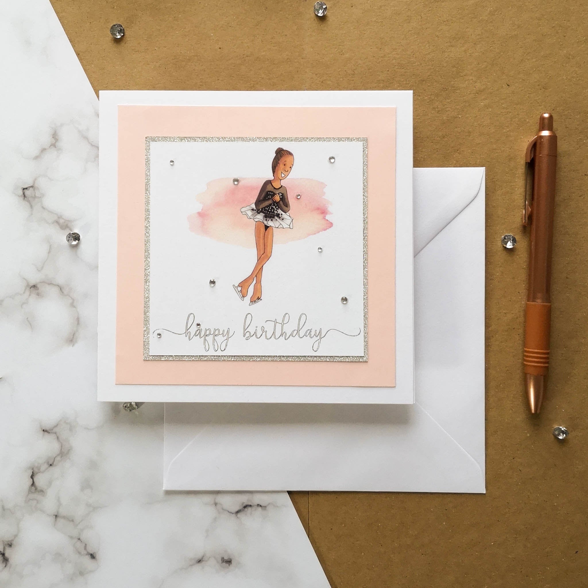 6 x 6 inch birthday card with image of a figure skater mid-flight, wearing a black dress. Pink watercolour brush stroke behind the figure skater illustration, with a silver foiled happy birthday script greeting and surrounded by clear/silver rhinestone gems